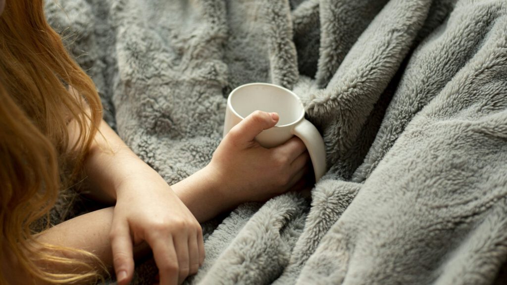 Eine Person sitzt in eine flauschige Decke gehüllt und hält eine Tasse