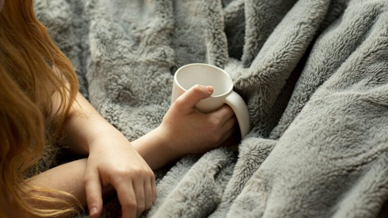 Eine Person sitzt in eine flauschige Decke gehüllt und hält eine Tasse