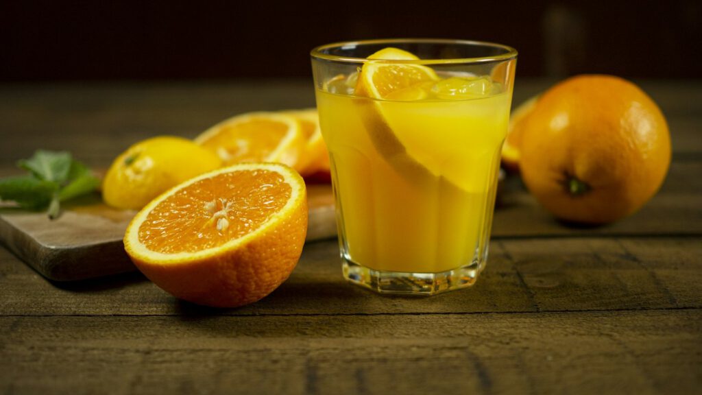Ein Glas frisch gepresster Saft, daneben halbierte Orangen und eine Zitrone auf einem Holztisch