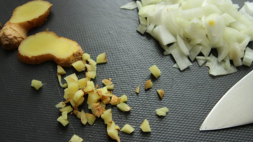 Ingwerscheiben, fein gehackter Ingwer und Zwiebelwürfel auf einem dunklen Schneidebrett, daneben eine Messerspitze