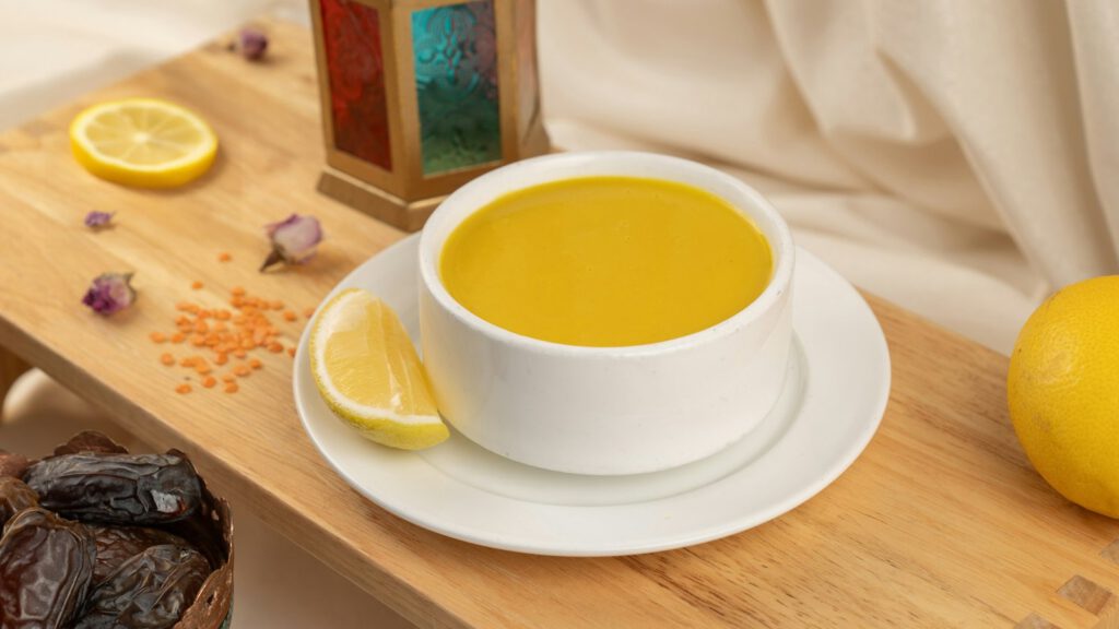 Eine Tasse mit gelber Suppe auf einem Holzbrett, daneben Zitronen und rote Linsen