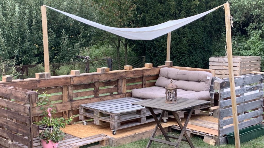 Selbst gebaute Sitzecke aus Holzpaletten mit Sonnensegel, Klapptisch und Pflanzen auf einer Wiese